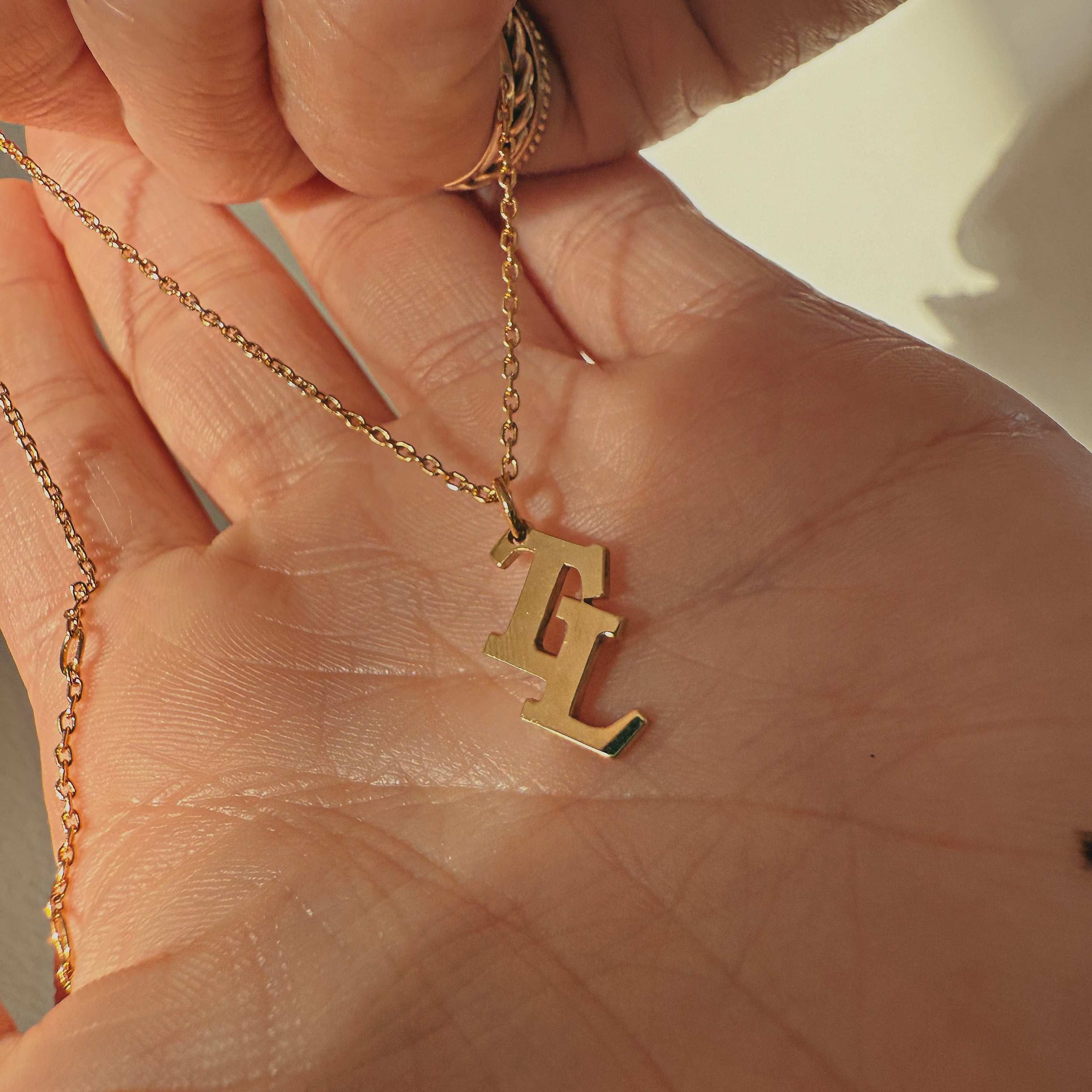 Gold necklace with a 'TL' pendant held in a hand against a neutral background